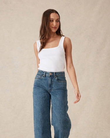 Woman wearing a white tank top and blue jeans against a beige background